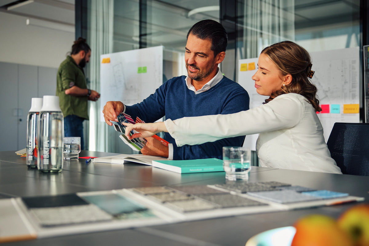 Two people working together at a desk with office supplies and a whiteboard in the background.